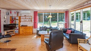 Gemütliches Interieur einer Hütte mit Holzbalkendecke, einer Couch, einem Couchtisch, einem Fernseher, einem Bücherregal und großen Fenstern mit Blick ins Freie.