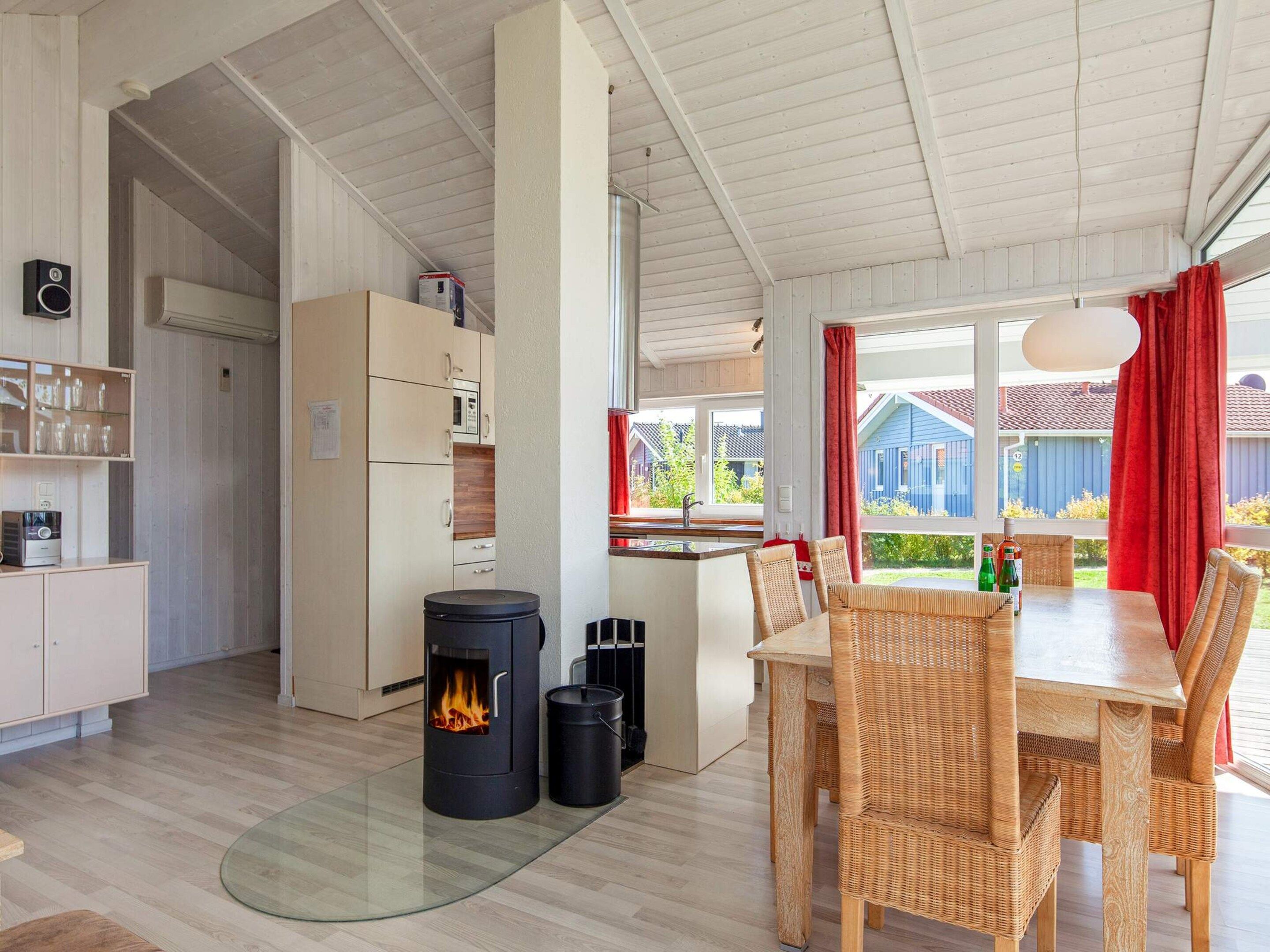 A modern kitchen with a wood-burning stove, a dining area with wicker chairs, and a view of a blue house through large windows.