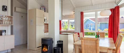 A modern kitchen with a wood-burning stove, a dining area with wicker chairs, and a view of a blue house through large windows.