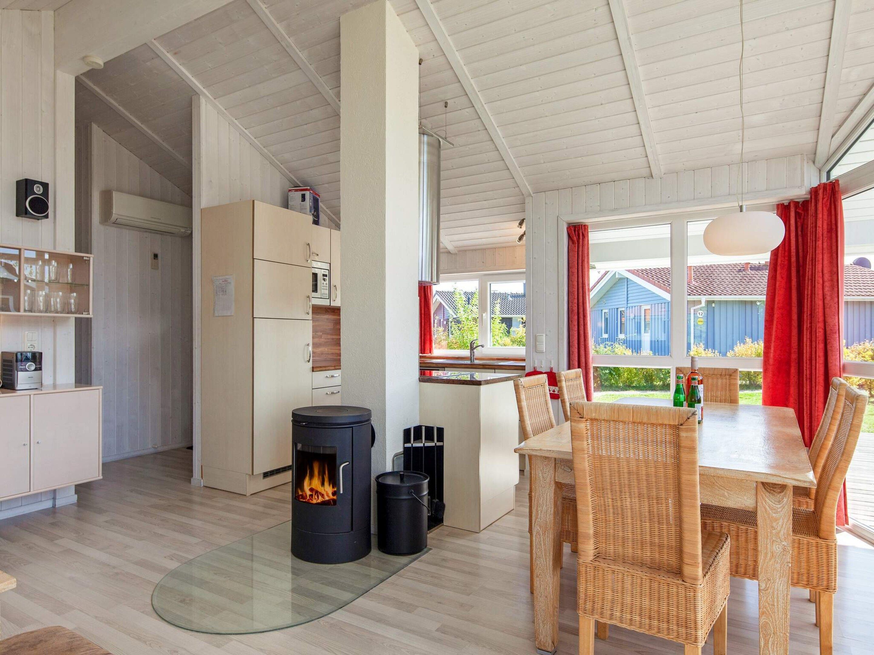 A modern kitchen with a wood-burning stove, a dining area with wicker chairs, and a view of a blue house through the window.