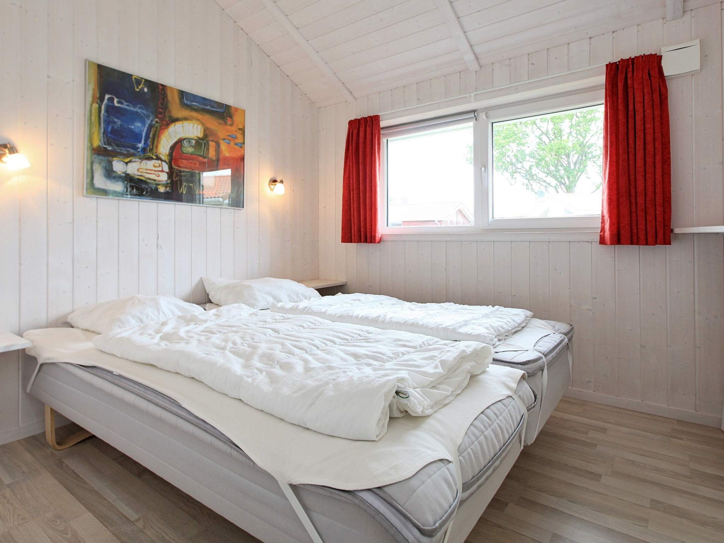A room with two beds, a window with red curtains, a painting on the wall, and a light fixture.