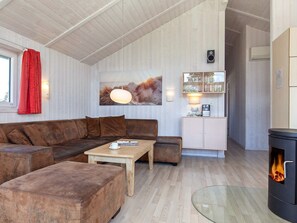 Uma sala de estar acolhedora com sofá, pufes, mesa de centro, mesa com tampo de vidro, lareira e área de cozinha com armários e luminária pendente.
