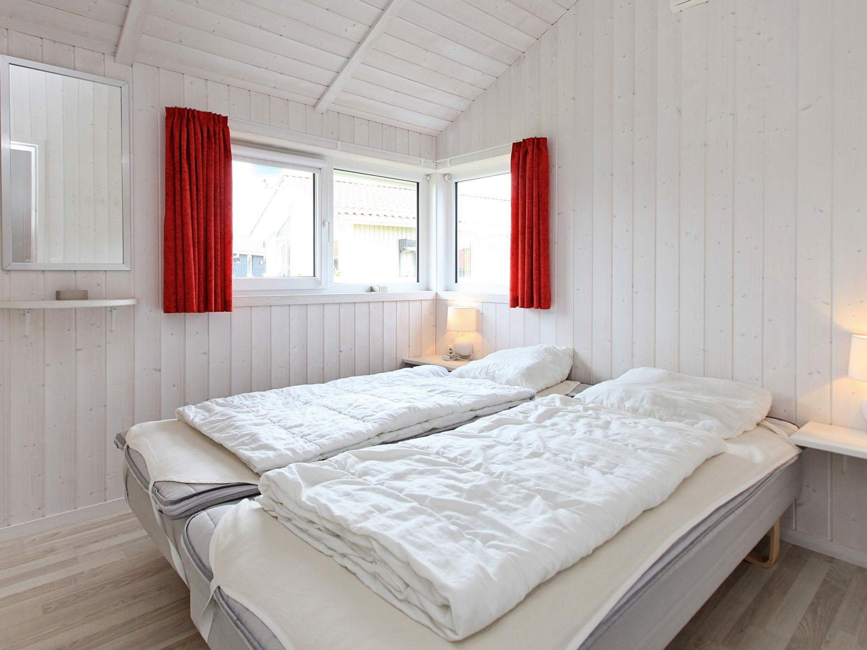 A room with two beds, a window with red curtains, a bedside table with a lamp, and a mirror on the wall.