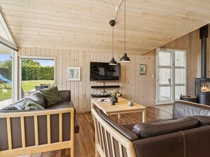 A cozy living room with wooden walls, a TV, a fireplace, and a large window overlooking a garden.