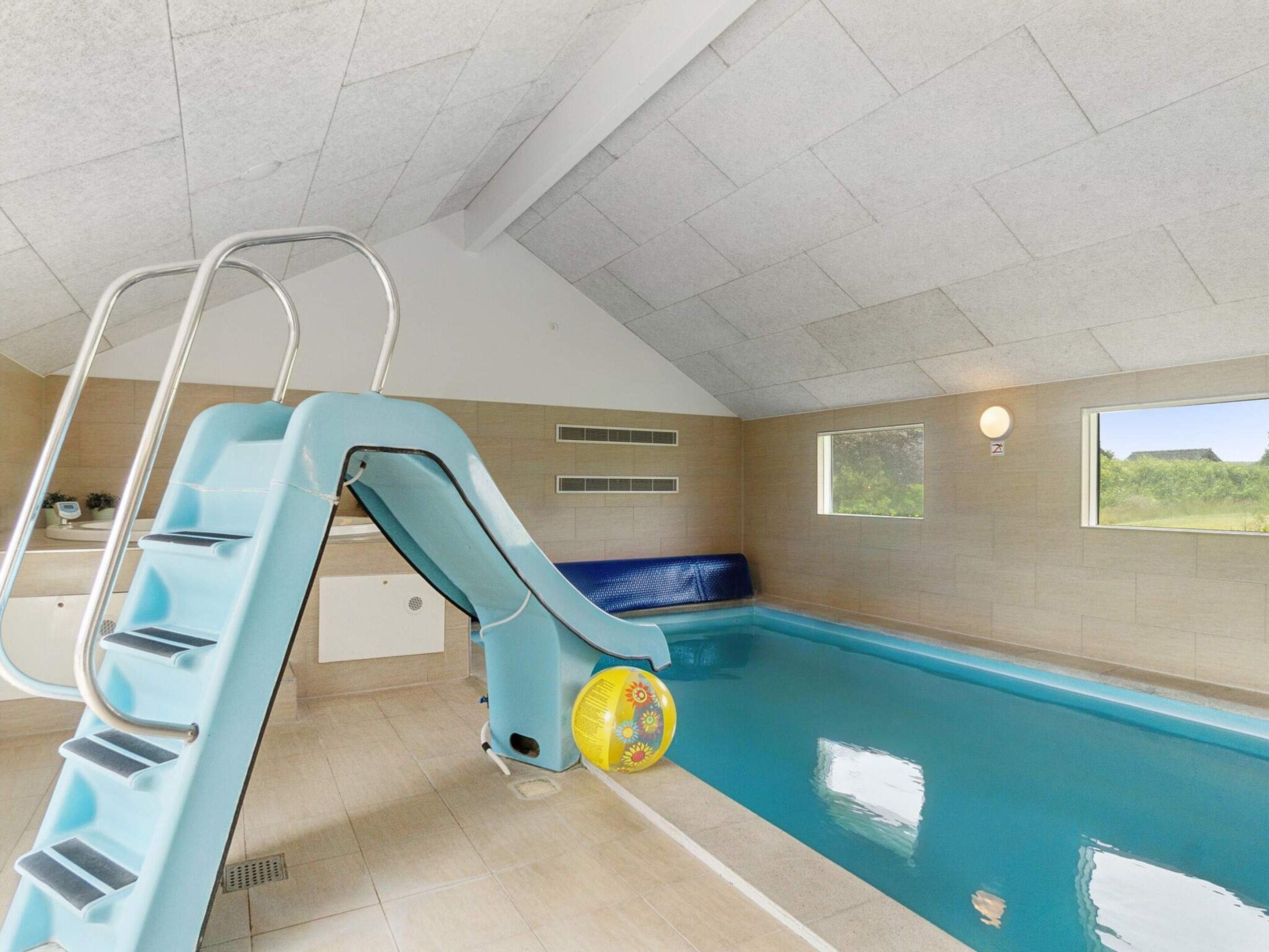 An indoor swimming pool with a blue slide, a sun lounger, and a view of greenery outside.
