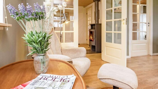 Un salon chaleureux avec une table basse en bois, un fauteuil confortable et un canapé. Un vase rempli de fleurs et un magazine sont posés sur la table.
