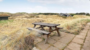 Une table et un banc de pique-nique en bois, installés sur une surface pavée, avec de l’herbe sèche et des dunes en arrière-plan.