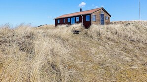 Une petite maison en bois d’un seul étage, avec un toit recouvert de tuiles rouges, entourée d’herbes hautes sur une colline.