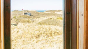 Une vue à travers la fenêtre d’un bâtiment, avec un paysage sablonneux et une petite construction visible au loin.