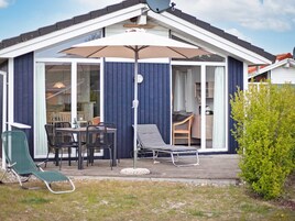 A covered outdoor seating area with a table and chairs, a lounge chair, and a small garden.