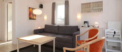 A modern living room with a sofa, a table, and a chair. There is a window with curtains, a radiator, and a shelf with glassware.