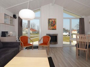 A modern living room with a sofa, chairs, a dining table, a fireplace, and a large window with a view of houses. - Holiday Home in GrÃ¶mmitz (Groemitz)