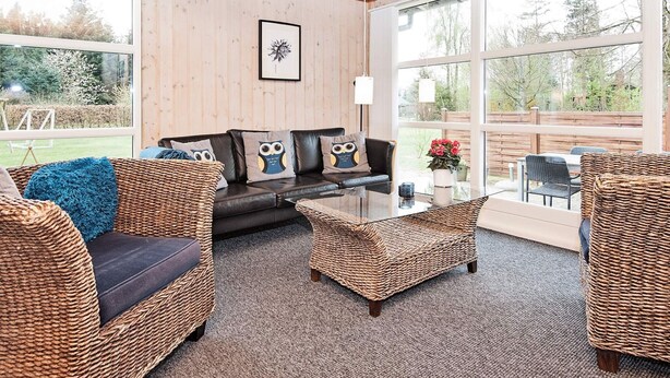 A cozy living room with wicker furniture, a dark leather sofa with owl-themed cushions, and large windows offering a view of the outdoors. - 5 Person Holiday Home in Ans By-by Traum (Ans)