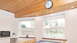 A room with a wooden ceiling, a dining table with chairs, a clock, and two windows with views of trees and a fence. - 5 Person Holiday Home in Ans By-by Traum (Ans)