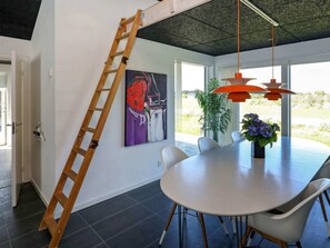 A dining area with a round table, chairs, a ladder, a painting, and a view through large windows.