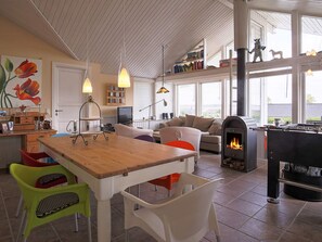 A cozy living room with a fireplace, a dining area with a wooden table, and a kitchen area with a bookshelf and a painting. - 5 Star Holiday Home in Jaegerspris (Jægerspris)