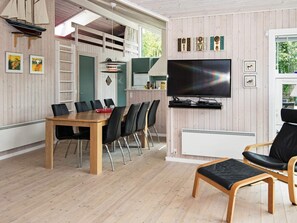A modern living room with a wooden floor, a dining area with a table and chairs, a television mounted on the wall, and a window with a view of greenery.