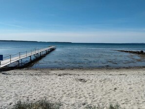 En strand med en träbrygga som sträcker sig ut i vattnet, en sandstrand och en klar himmel.