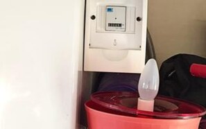Un récipient rouge doté d’un couvercle, contenant un objet blanc à l’intérieur, peut-être un ustensile, dans une pièce dont le mur et la porte sont blancs.