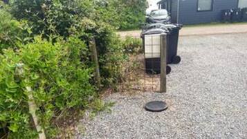 A gravel driveway with a metal fence, a black trash bin, and a blue building in the background.