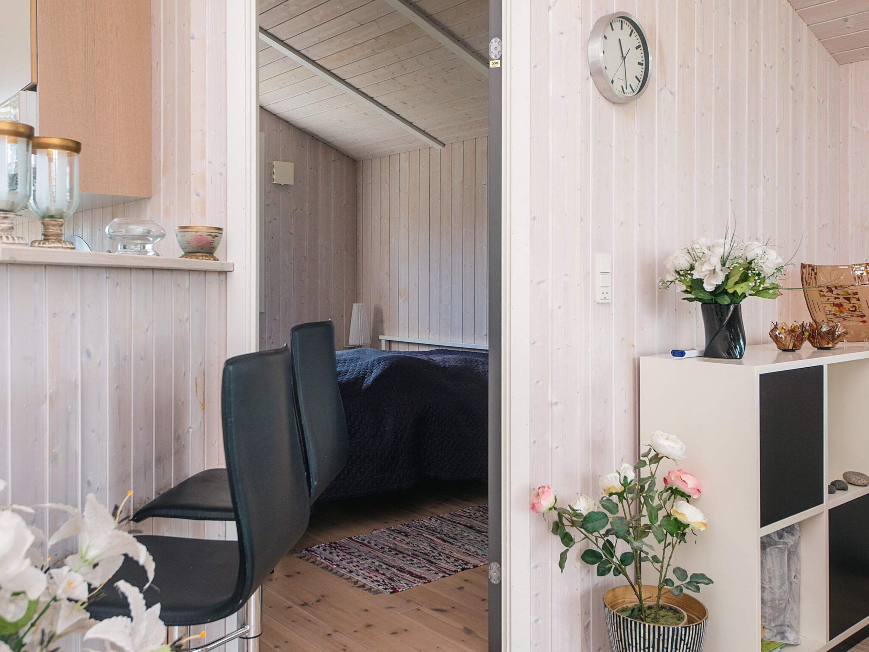 Una habitación con cama, una silla, un escritorio con computadora, un reloj y un florero con flores.