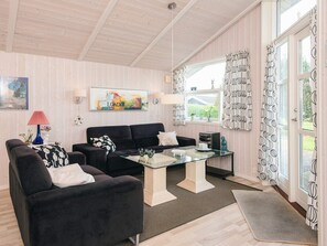 A living room with a sofa, a glass coffee table, and a TV. There is a window with curtains and a painting on the wall.