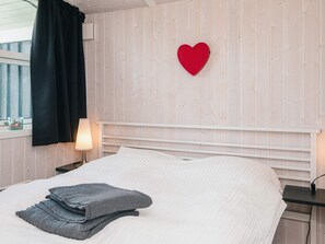 Ein Bett mit weißem Kopfteil, eine Wanddekoration in Herzform in Rot, eine Lampe auf einem Nachttisch und ein Fenster mit Vorhängen.