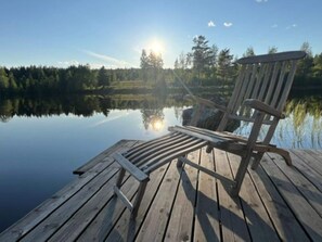En träbrygga med en stol som vetter mot en stilla sjö, omgiven av träd och en klar himmel.