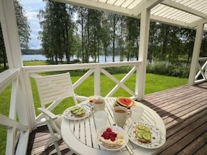 Ett takförsett utomhusområde med sittplatser och ett bord dukat för frukost, med utsikt över en sjö och omgivet av träd.