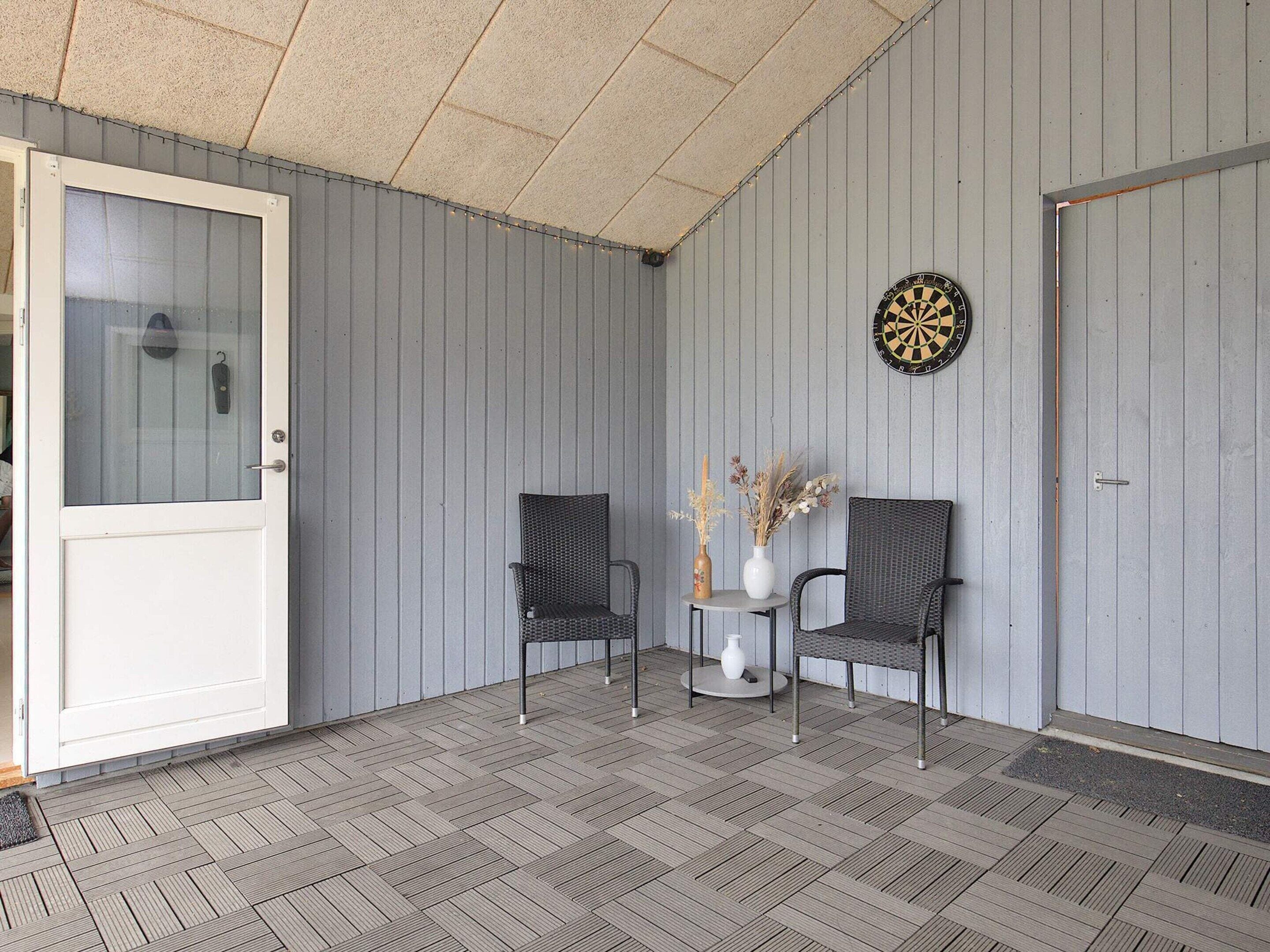 A covered porch with two wicker chairs, a small table with a vase, and a dartboard on the wall.