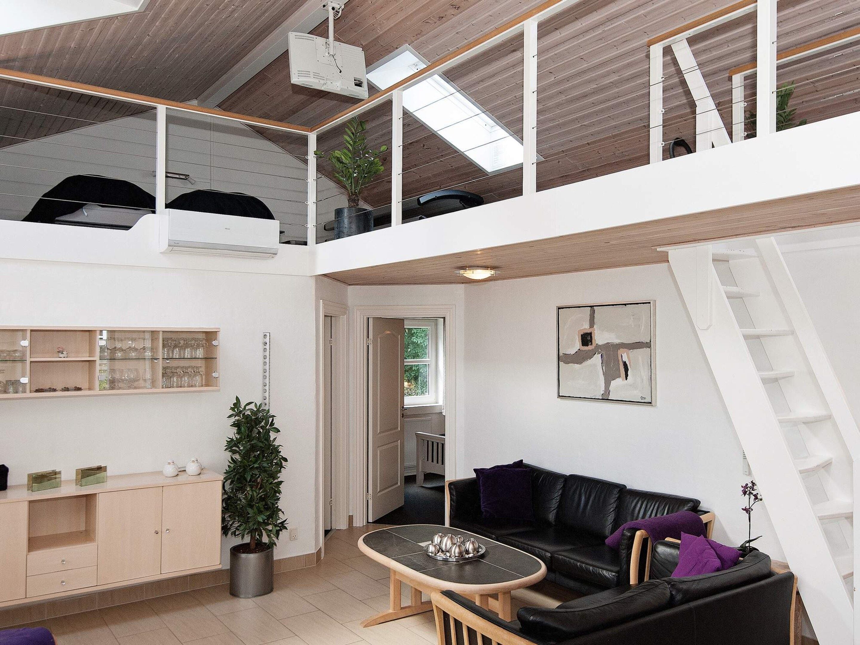 A spacious loft-style living area with a high ceiling, a mezzanine level, a black leather sofa, a coffee table, and a potted plant.