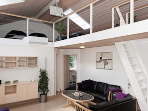 Amplo espaço de estar estilo loft com pé direito alto, mezanino, sofá de couro preto, mesa de centro e planta em vaso.