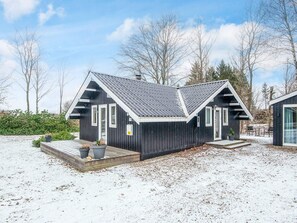 A modern cabin with a dark exterior, white trim, and a steeply pitched roof. It features a front porch with steps and potted plants, and is surrounded by a snowy landscape with trees in the background. - 6 Person Holiday Home in Toftlund (Toftlund)
