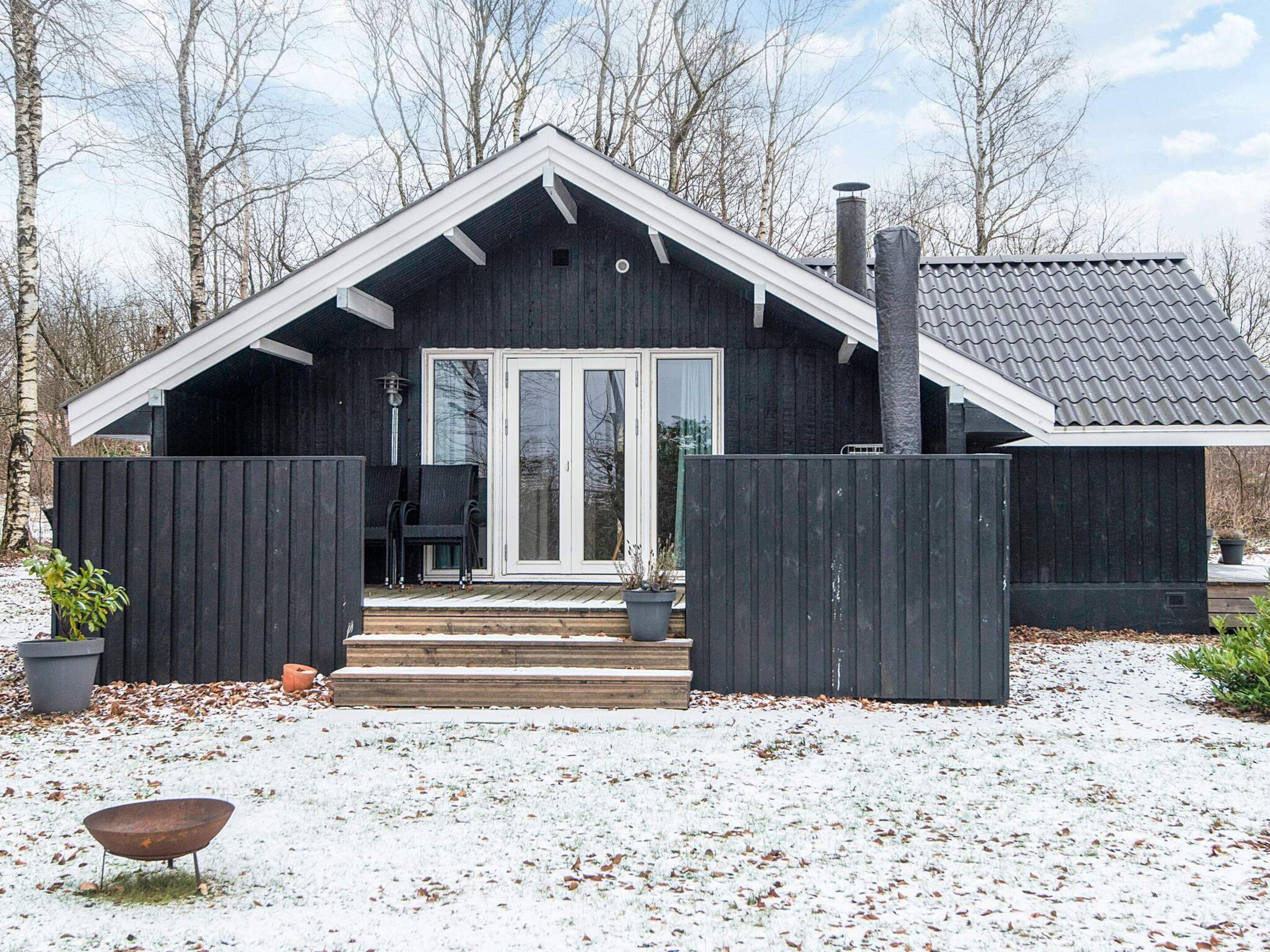A wooden cabin with a dark exterior, white trim, and a chimney. There is a covered porch with steps leading to a door, and a small outdoor seating area with chairs.