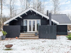 A wooden cabin with a dark exterior, white trim, and a chimney. There is a covered porch with steps leading to a door, and a small outdoor seating area with chairs. - 6 Person Holiday Home in Toftlund (Toftlund)