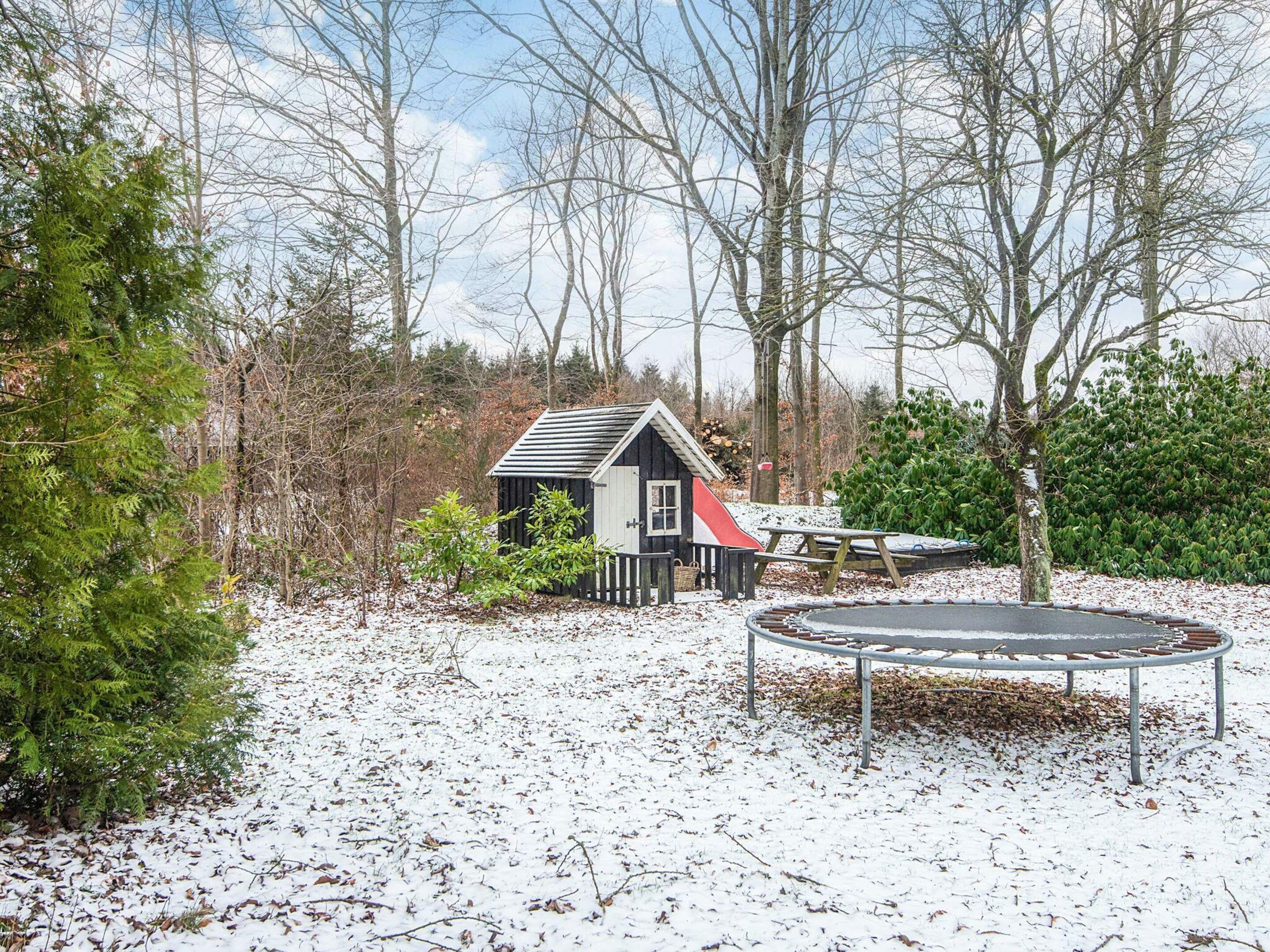 Een klein houten huisje met een schuin dak, omringd door bomen en een trampoline in een besneewd landschap.