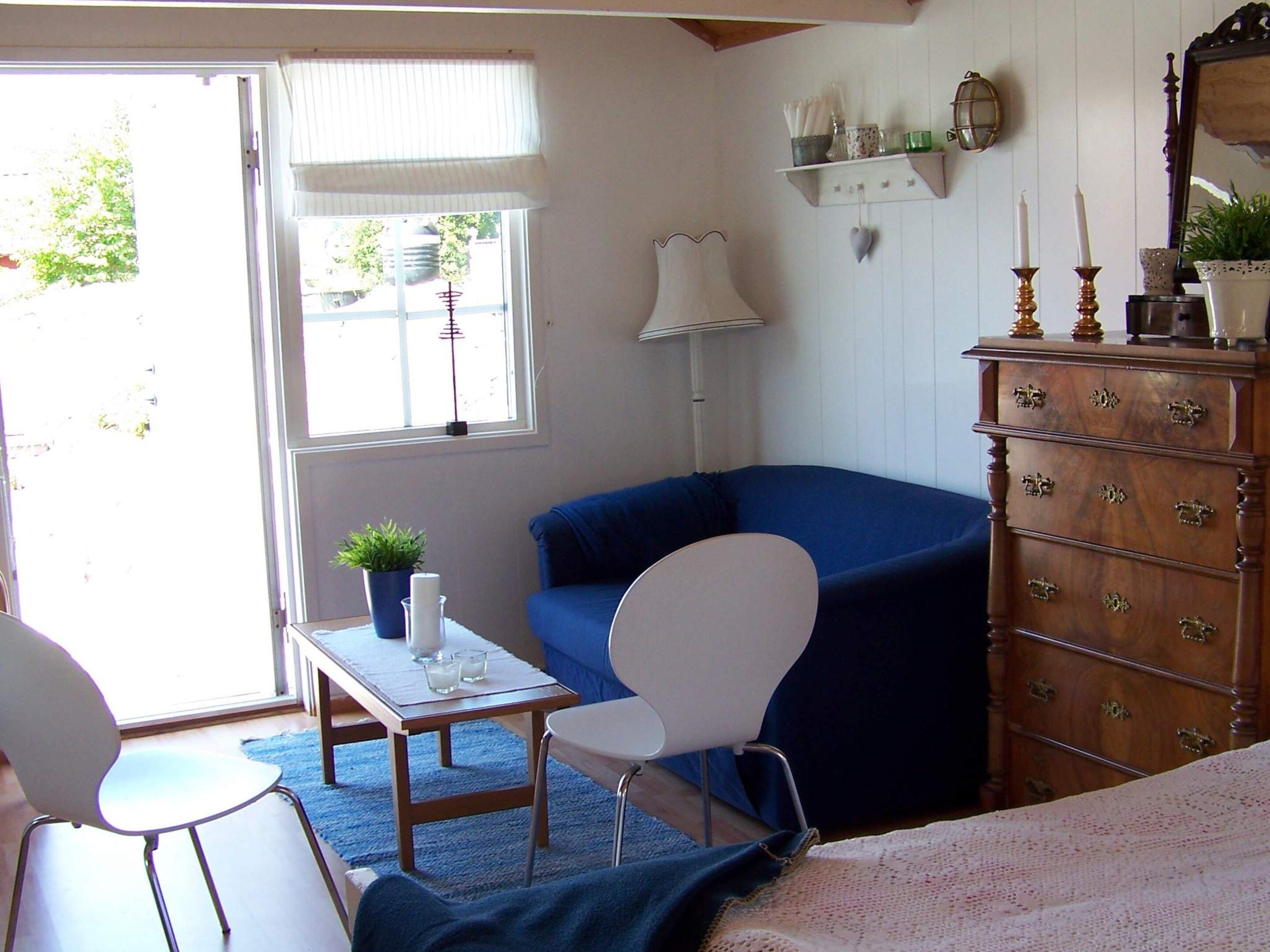 A room with a blue armchair, a white chair, a wooden dresser, and a small table with a plant and candles.