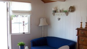 Quarto com sofá azul, cadeira branca, mesa de madeira com planta, abajur e cômoda com gavetas.