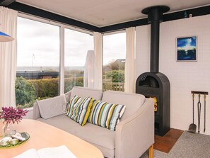 Ein gemütliches Wohnzimmer mit einer Couch, einem Tisch mit einer Vase und einem Buch, einem Kamin und Blick ins Freie.