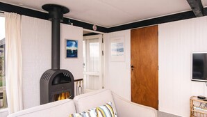 A room with a white brick wall, a black wood-burning stove, a white sofa with striped cushions, and a television on a wooden stand. - 6 Person Holiday Home in Borkop (Borkop)