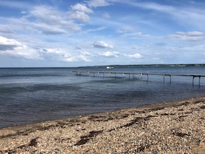 A pebbled beach with a wooden pier extending into calm waters, a boat in the distance, and a cloudy sky.