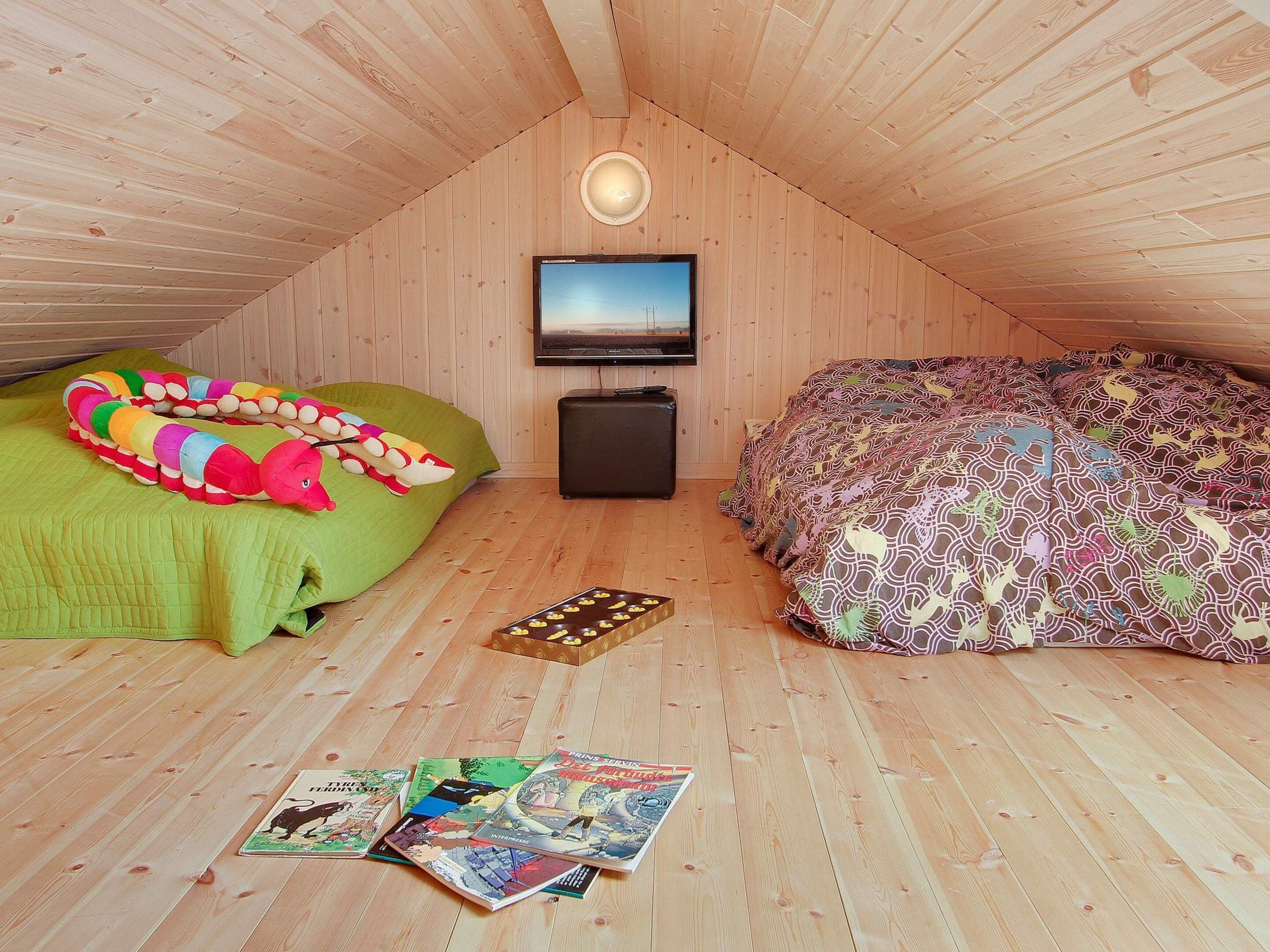 A cozy attic bedroom with a sloped ceiling, a flat-screen TV mounted on the wall, a green and a patterned bed, and a small chest of drawers.