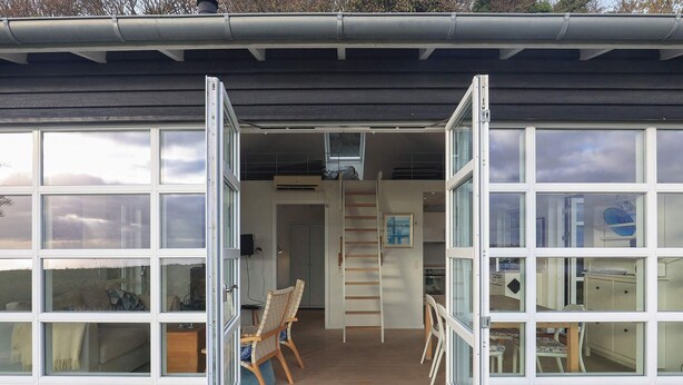 A modern house with large windows, a dining area with chairs, a ladder, and a kitchen visible through the open doors.