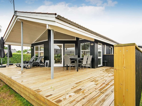Uma área externa coberta com deck de madeira, espreguiçadeiras e uma mesa com cadeiras.
