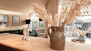 Un interior de madera con un ventanal grande, una mesa de madera, un jarrón con plantas secas y una silla.
