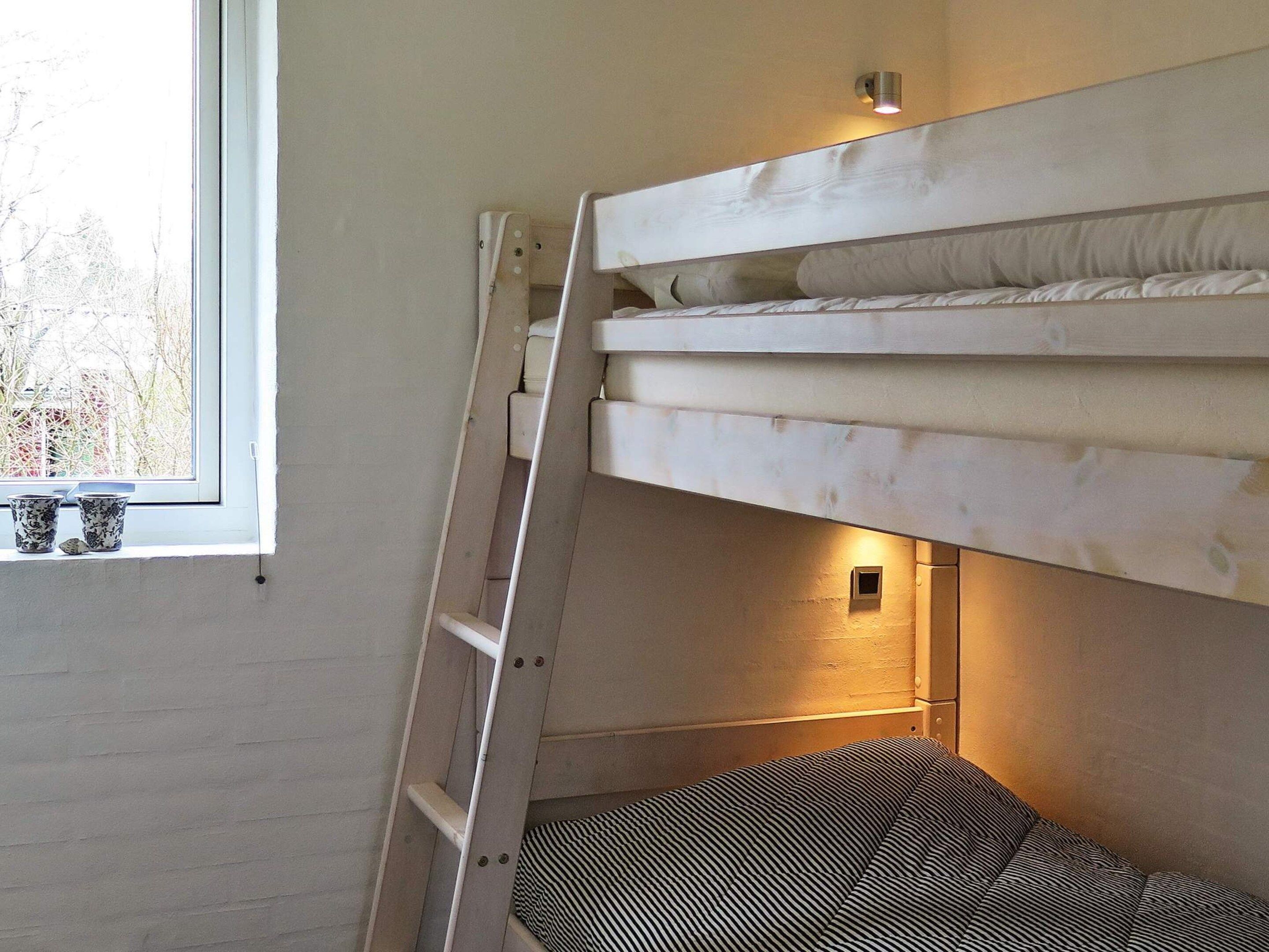 A bunk bed with a ladder, a window with two cups on the sill, and a wall-mounted light.