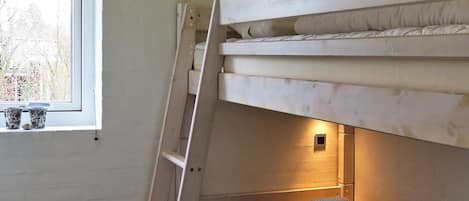 A bunk bed with a ladder, a window with two cups on the sill, and a wall-mounted light.