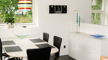 A modern dining area with a white table, black chairs, and a glass bowl. There is a window with a view of greenery outside.