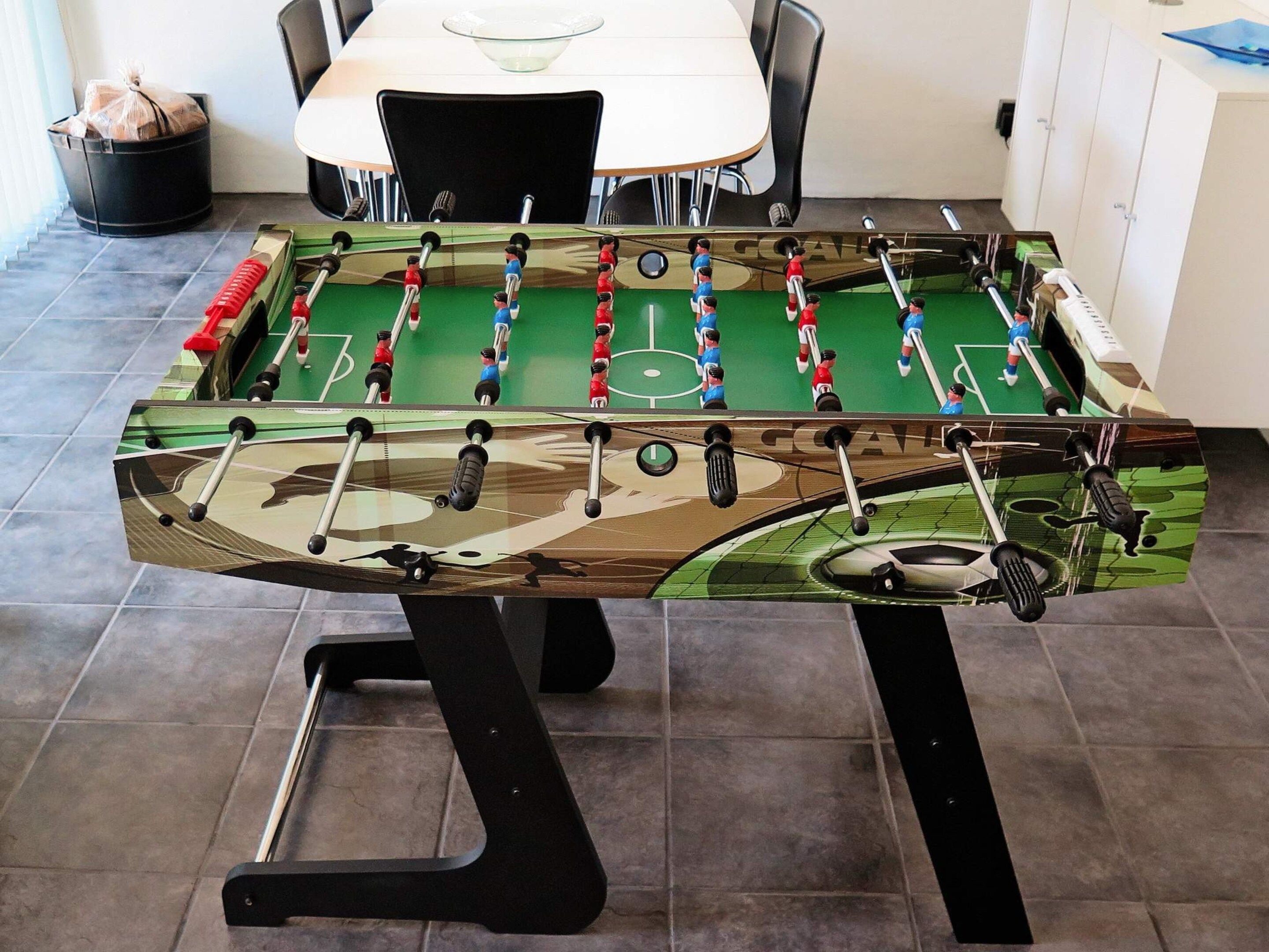 Una mesa de fútbolín con diseño de camuflaje, ubicada en una habitación con suelo de baldosas y una mesa blanca al fondo.