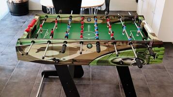 A foosball table with a camouflage design, positioned in a room with a tiled floor and a white table in the background.